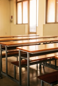 An empty classroom with wooden desks and benches arranged in rows. Large windows allow natural light to fill the room, creating a warm and somewhat nostalgic atmosphere. The room appears clean and well-maintained, with tiled floors and an organized layout.