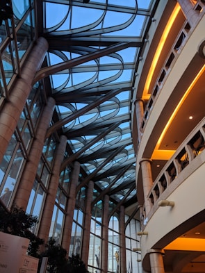 A modern architectural interior featuring a large atrium with a glass ceiling. Arched metal supports extend across the roof, creating a geometric pattern against the blue sky. Tall stone columns line the sides, supporting multiple levels with rounded balconies. Warm lighting highlights the interiors of the balconies.