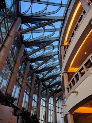 A modern architectural interior featuring a large atrium with a glass ceiling. Arched metal supports extend across the roof, creating a geometric pattern against the blue sky. Tall stone columns line the sides, supporting multiple levels with rounded balconies. Warm lighting highlights the interiors of the balconies.