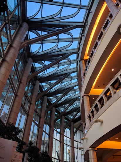 A modern architectural interior featuring a large atrium with a glass ceiling. Arched metal supports extend across the roof, creating a geometric pattern against the blue sky. Tall stone columns line the sides, supporting multiple levels with rounded balconies. Warm lighting highlights the interiors of the balconies.