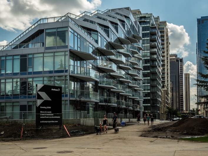 A modern multi-story glass building with balconies extends along an urban street, displaying a sign about upcoming condominium availability. Several people are walking on the adjacent sidewalk, some with dogs, and new construction seems underway. The sky is partly cloudy, and surrounding buildings indicate a dense city setting.