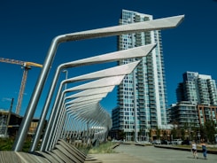 A series of modern architectural structures with metallic beams align in a geometric pattern. High-rise residential buildings stand in the background against a deep blue sky. A crane is visible to the left, and a boardwalk with grass and people can be seen in the foreground.