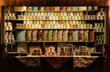 A wooden shop display filled with intricately patterned and colorful ceramic mugs on shelves. Various designs featuring floral and geometric patterns are visible. The lower shelves show similar patterns on rectangular objects. A person is partially visible behind the counter.