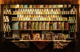 A wooden shop display filled with intricately patterned and colorful ceramic mugs on shelves. Various designs featuring floral and geometric patterns are visible. The lower shelves show similar patterns on rectangular objects. A person is partially visible behind the counter.