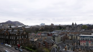 Aerial view of a historic cityscape with a blend of old and modern architecture. The foreground features stone buildings with intricate designs and spires. In the background, rolling hills and cloudy skies provide a natural backdrop. Streets are visible with parked cars, adding an urban touch.