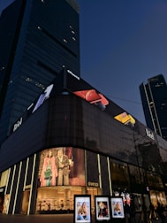 A tall, modern building with a large electronic billboard displaying an iPhone advertisement at the top. Below, there's a Gucci storefront with illuminated logo signs, showcasing fashion imagery on digital screens. The scene is set during the evening with a clear sky in the background.