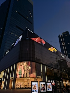 A tall, modern building with a large electronic billboard displaying an iPhone advertisement at the top. Below, there's a Gucci storefront with illuminated logo signs, showcasing fashion imagery on digital screens. The scene is set during the evening with a clear sky in the background.