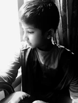 A serene moment of a child in a cream kurta set, standing by a window with gentle sunlight casting warm shadows.
