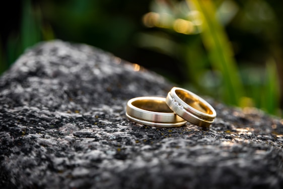 Two gold wedding rings are placed on a textured gray rock surface, with a blurred natural green background. One ring is plain, while the other is adorned with small diamonds.