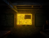 A bright neon sign displays the words 'Stay Hydrated' against a dark brick wall, with additional chalkboard-style menu signs on either side. The surrounding area appears dimly lit, with a subtle presence of a person to the right sitting at a table.