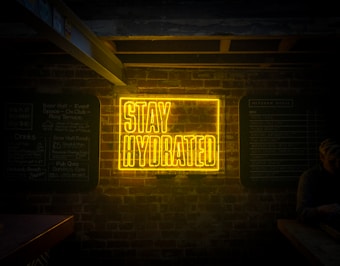 A bright neon sign displays the words 'Stay Hydrated' against a dark brick wall, with additional chalkboard-style menu signs on either side. The surrounding area appears dimly lit, with a subtle presence of a person to the right sitting at a table.