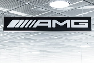 A black sign displays the letters 'AMG' in bold white font. Above it is a metallic, grid-patterned ceiling with modern recessed lighting fixtures. The scene gives an industrial and sleek aesthetic.