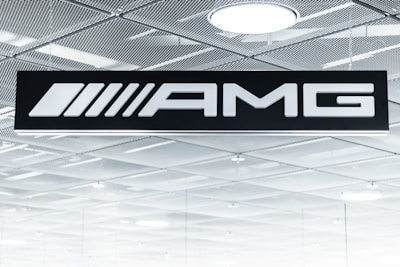 A black sign displays the letters 'AMG' in bold white font. Above it is a metallic, grid-patterned ceiling with modern recessed lighting fixtures. The scene gives an industrial and sleek aesthetic.