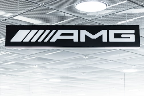 A black sign displays the letters 'AMG' in bold white font. Above it is a metallic, grid-patterned ceiling with modern recessed lighting fixtures. The scene gives an industrial and sleek aesthetic.