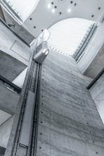 A view from inside an industrial building showcases a tall, gray concrete elevator shaft reaching upwards. A modern, minimalistic elevator car travels along the vertical tracks. The lighting is soft and cool, highlighting the sleek, functional design of the architecture. The high ceiling is curved with embedded lights.