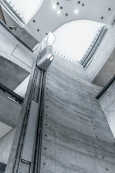 A view from inside an industrial building showcases a tall, gray concrete elevator shaft reaching upwards. A modern, minimalistic elevator car travels along the vertical tracks. The lighting is soft and cool, highlighting the sleek, functional design of the architecture. The high ceiling is curved with embedded lights.