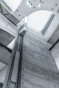 A view from inside an industrial building showcases a tall, gray concrete elevator shaft reaching upwards. A modern, minimalistic elevator car travels along the vertical tracks. The lighting is soft and cool, highlighting the sleek, functional design of the architecture. The high ceiling is curved with embedded lights.
