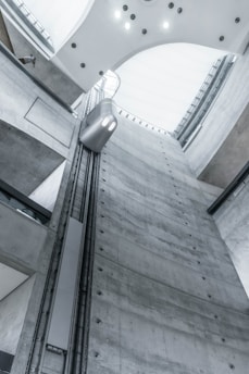 A view from inside an industrial building showcases a tall, gray concrete elevator shaft reaching upwards. A modern, minimalistic elevator car travels along the vertical tracks. The lighting is soft and cool, highlighting the sleek, functional design of the architecture. The high ceiling is curved with embedded lights.