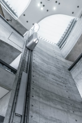 A view from inside an industrial building showcases a tall, gray concrete elevator shaft reaching upwards. A modern, minimalistic elevator car travels along the vertical tracks. The lighting is soft and cool, highlighting the sleek, functional design of the architecture. The high ceiling is curved with embedded lights.