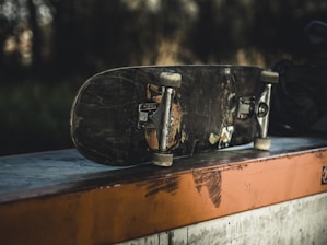 A worn skateboard with visible scratches and chipped paint rests upside down on a concrete ledge. The background is blurred, giving a sense of focus on the skateboard. Sunlight casts shadows, creating a gritty, urban feel.