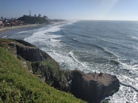 Tall cliffs with lush greenery overlook a vast ocean, with white waves crashing gently along the shoreline. A couple of people stand on the edge of the cliff, enjoying the panoramic view of the sea. In the distance, a beach with people enjoying the day stretches along the coast, with buildings and a hill with communication towers visible in the background.