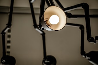 A close-up view of an industrial-style lamp with an exposed light bulb. The lamp has a metallic structure with multiple adjustable arms, creating a mechanical and modern appearance. The lighting creates a contrast against the muted background.