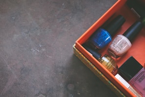 A collection of nail polish bottles arranged neatly inside an orange box. The box is partially open, revealing various shades including blue, beige, yellow, and pink. The box is placed on a textured, dark surface, possibly a tabletop or floor.
