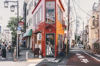 A vibrant urban street scene featuring a narrow, brightly colored red building at a corner. The streets are lined with buildings and power lines, and a few pedestrians are walking, including a person with a bicycle. Signs and storefronts suggest a bustling, lively atmosphere.