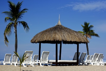 A tranquil beach scene featuring a thatched-roof gazebo surrounded by white lounge chairs. Two palm trees stand nearby, under a clear blue sky. The sandy beach and calm ocean provide a peaceful backdrop.