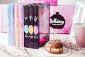 A collection of colorful books is displayed in a row next to a decorative mug and a donut on a lace doily. The background features a blurred pink and black design with a framed sign. The books have eye-catching pastel and dark covers with intricate titles.