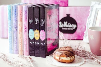 A collection of colorful books is displayed in a row next to a decorative mug and a donut on a lace doily. The background features a blurred pink and black design with a framed sign. The books have eye-catching pastel and dark covers with intricate titles.