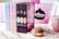 A collection of colorful books is displayed in a row next to a decorative mug and a donut on a lace doily. The background features a blurred pink and black design with a framed sign. The books have eye-catching pastel and dark covers with intricate titles.