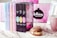 A collection of colorful books is displayed in a row next to a decorative mug and a donut on a lace doily. The background features a blurred pink and black design with a framed sign. The books have eye-catching pastel and dark covers with intricate titles.