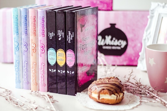 A collection of colorful books is displayed in a row next to a decorative mug and a donut on a lace doily. The background features a blurred pink and black design with a framed sign. The books have eye-catching pastel and dark covers with intricate titles.
