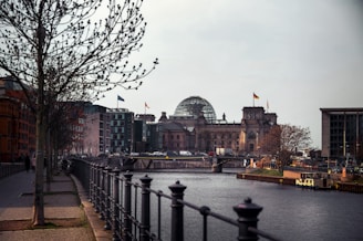 A river flows through an urban area with prominent buildings featuring modern and classic architecture; a notable dome and flags are visible on one of the buildings. Trees line the sidewalk along the river, with a black railing in the foreground and a stone walkway beside it. Traffic and pedestrians are present near the historic structure.