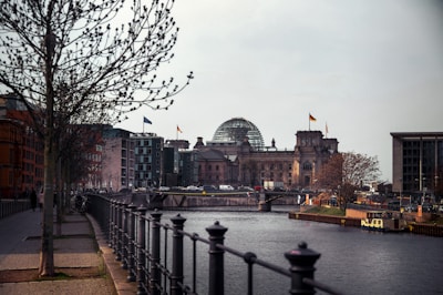 A river flows through an urban area with prominent buildings featuring modern and classic architecture; a notable dome and flags are visible on one of the buildings. Trees line the sidewalk along the river, with a black railing in the foreground and a stone walkway beside it. Traffic and pedestrians are present near the historic structure.