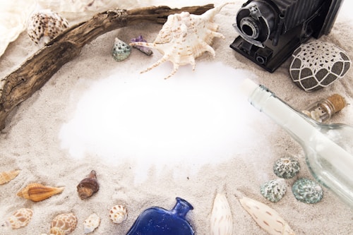 A collection of seashells, driftwood, and a vintage camera are arranged on a sandy surface. A glass bottle with a cork lies to the right, alongside a smaller bottle. There is a central area of white sand surrounded by various shells and decorative items.