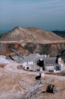 A large industrial quarry site featuring mounds of gravel and dirt, with several pieces of machinery positioned around the area. There are pathways through the site, piles of rock, and various metal structures.
