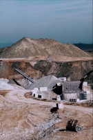 A large industrial quarry site featuring mounds of gravel and dirt, with several pieces of machinery positioned around the area. There are pathways through the site, piles of rock, and various metal structures.