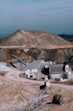 A large industrial quarry site featuring mounds of gravel and dirt, with several pieces of machinery positioned around the area. There are pathways through the site, piles of rock, and various metal structures.