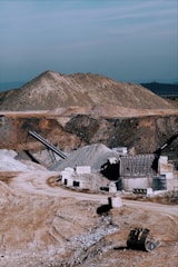 A large industrial quarry site featuring mounds of gravel and dirt, with several pieces of machinery positioned around the area. There are pathways through the site, piles of rock, and various metal structures.