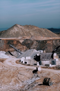 A large industrial quarry site featuring mounds of gravel and dirt, with several pieces of machinery positioned around the area. There are pathways through the site, piles of rock, and various metal structures.