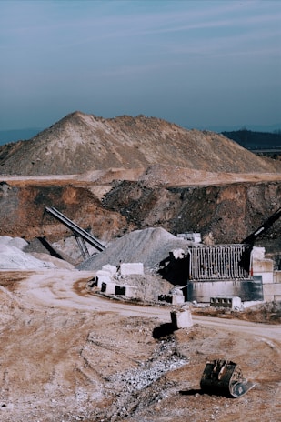 A large industrial quarry site featuring mounds of gravel and dirt, with several pieces of machinery positioned around the area. There are pathways through the site, piles of rock, and various metal structures.
