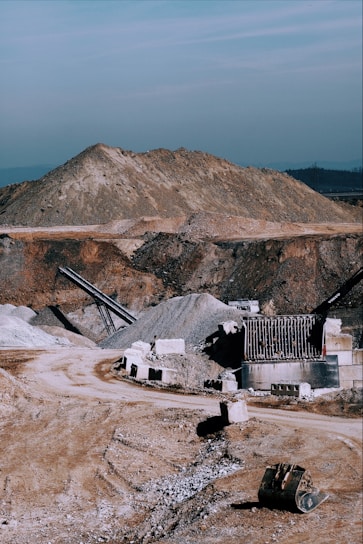 A large industrial quarry site featuring mounds of gravel and dirt, with several pieces of machinery positioned around the area. There are pathways through the site, piles of rock, and various metal structures.