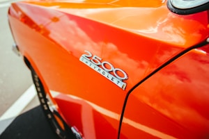 A close-up view of a classic Chevrolet 2500 badge on an orange car, with reflections of clouds on the glossy paint surface. The image captures the sleek design and vibrant color of the vehicle.