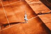 A tennis player is performing a serve on a clay court. The player's body is slightly tilted with one arm raised, and the shadow cast onto the court is prominent. The warm sunlight creates contrasting shadows and highlights across the court's surface.