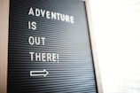 A letter board displays the motivational message 'Adventure is out there!' in white letters against a black background. Positioned diagonally, an arrow points to the right, suggesting movement or direction.