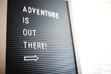 A letter board displays the motivational message 'Adventure is out there!' in white letters against a black background. Positioned diagonally, an arrow points to the right, suggesting movement or direction.