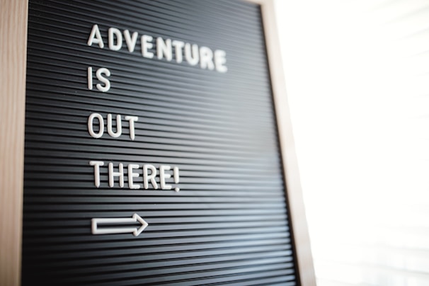 A letter board displays the motivational message 'Adventure is out there!' in white letters against a black background. Positioned diagonally, an arrow points to the right, suggesting movement or direction.