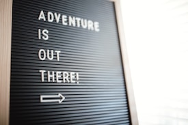 A letter board displays the motivational message 'Adventure is out there!' in white letters against a black background. Positioned diagonally, an arrow points to the right, suggesting movement or direction.